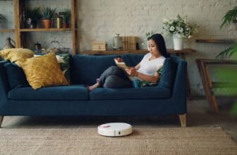 Woman reading on sofa while a robot vacuum cleans the floor in a cozy living room.