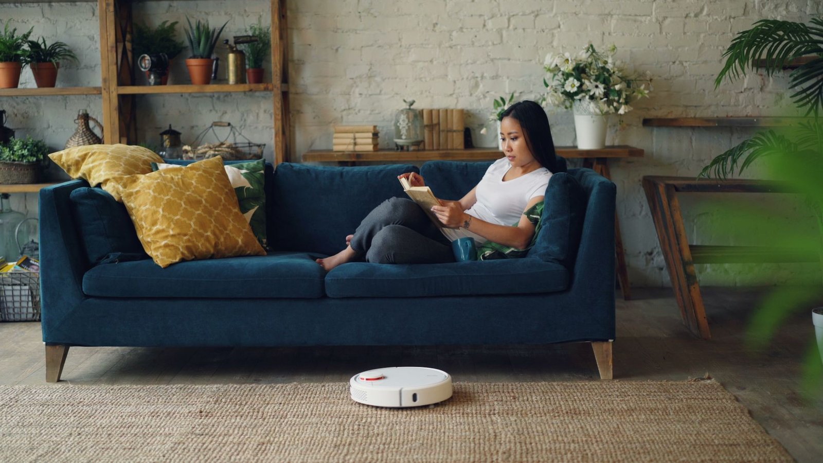 Woman reading on sofa while a robot vacuum cleans the floor in a cozy living room.