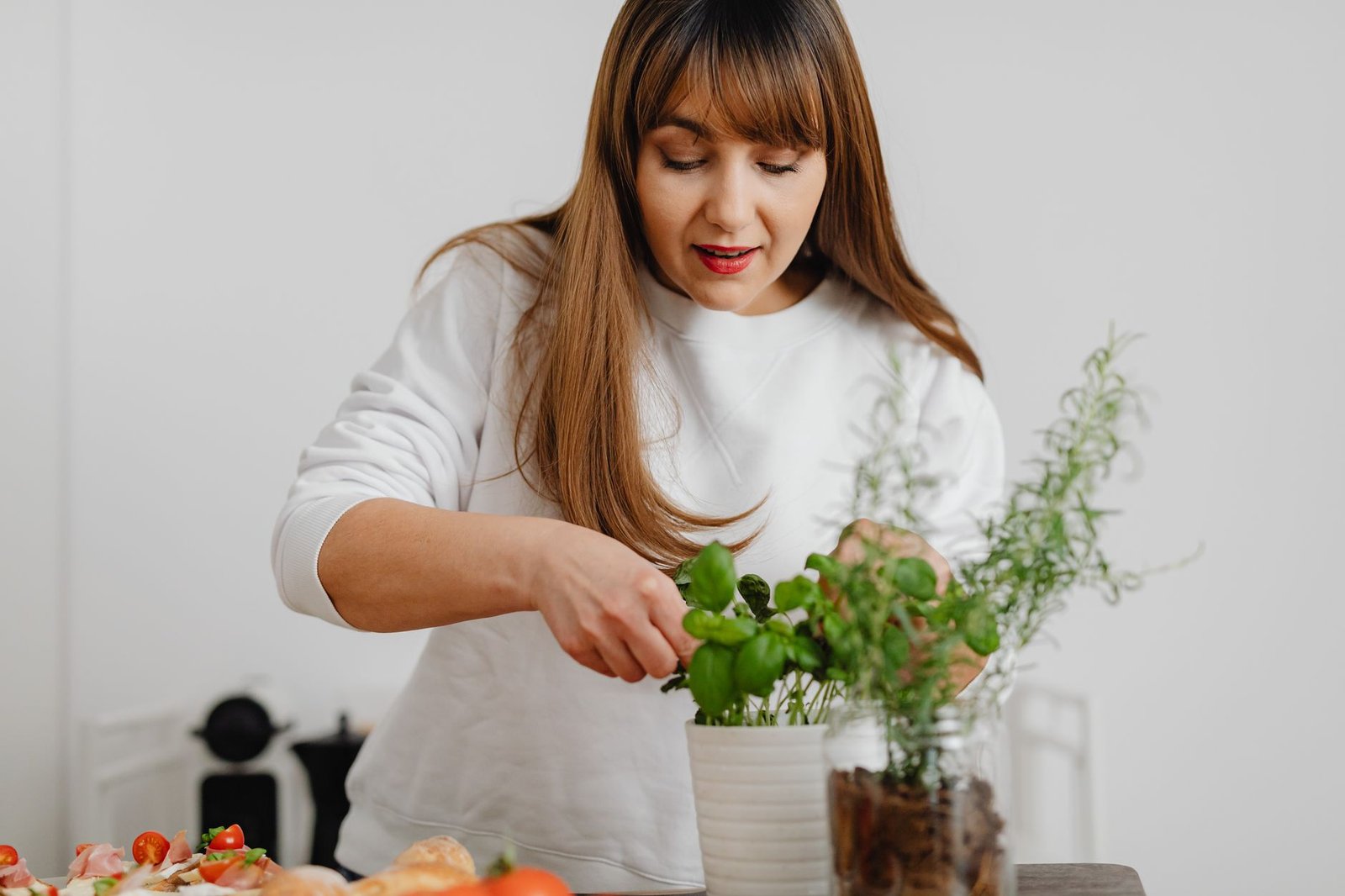 A woman in a white sweater tending to a potted herb plant indoors, showing focus and care.