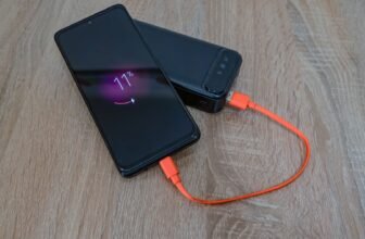 A smartphone charging from a power bank with an orange cable on a wooden table.