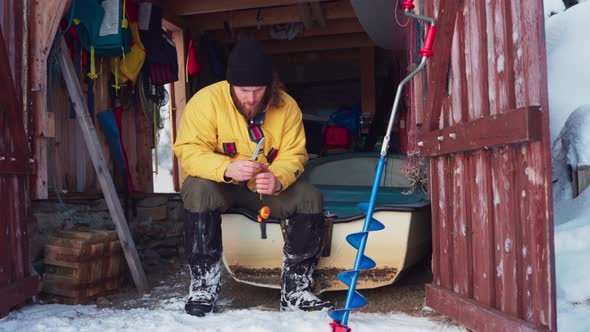 Man In Wooden Storage Sits On A Boat While Preparing Gear For Ice Fishing In Winter. full shot