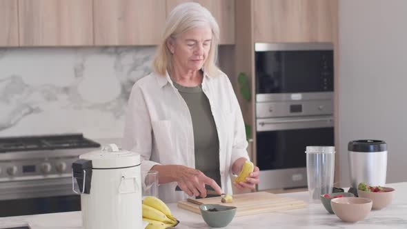 Senior woman grabbing banana, slicing and peeling at marble counter, adding to blender for smoothie