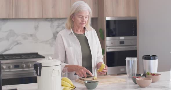 Senior woman grabbing banana, slicing and peeling at marble counter, adding to blender for smoothie
