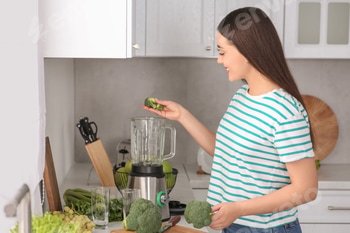 Woman adding broccoli into blender for smoothie in kitchen